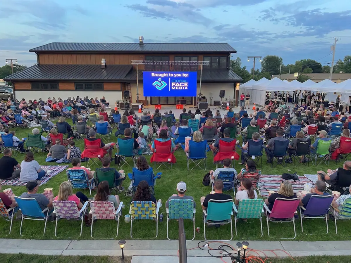 Families in lawn chairs watching a City Park Events video wall in front of a community building at dusk